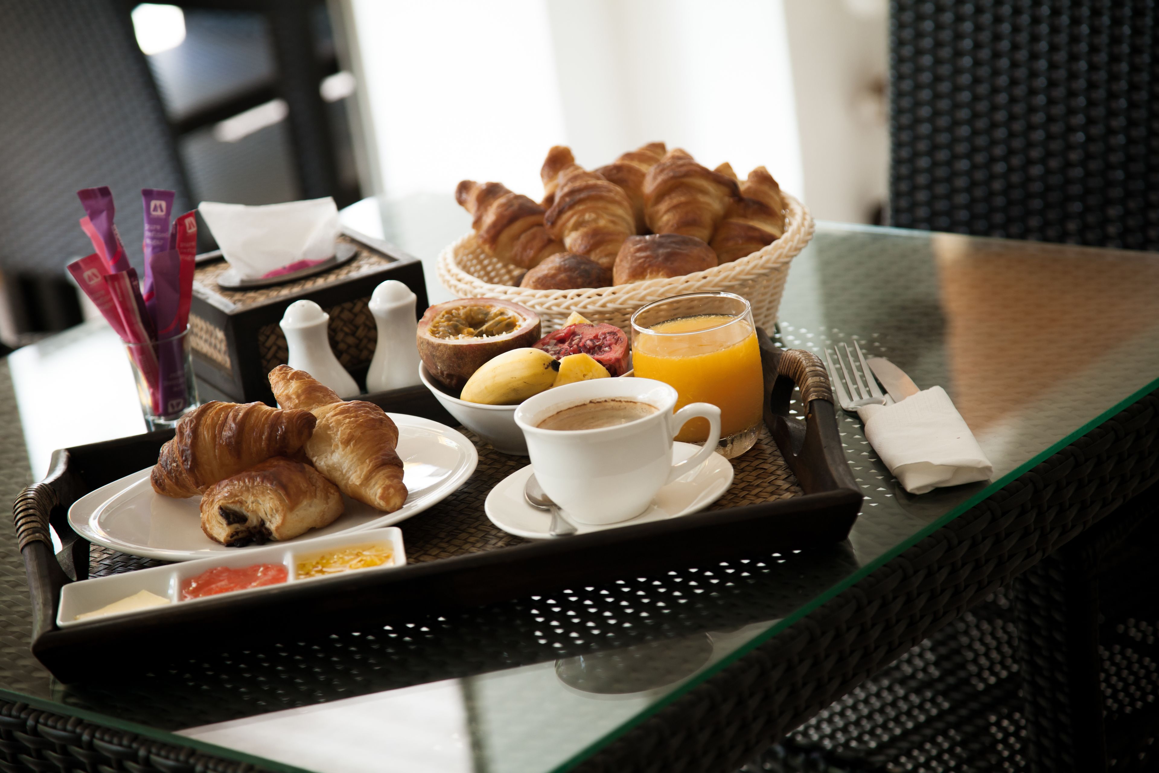 Ein Frühstücksbrett mit Croissants, Obst, Kaffee und Orangensaft auf einem Glastisch.