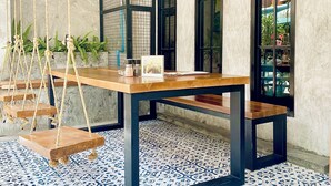 A room with a wooden table, black metal frame, and a patterned floor. There are two windows with black frames, a white ceiling with a grid pattern, and a hanging rope swing with plants. - BH Baan Paradise (Patong)