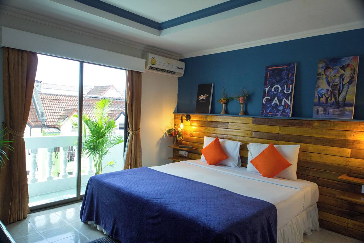 Habitación de hotel con una cama grande, cabecera de madera, balcón con vistas a edificios y una pared decorada con carteles motivacionales.