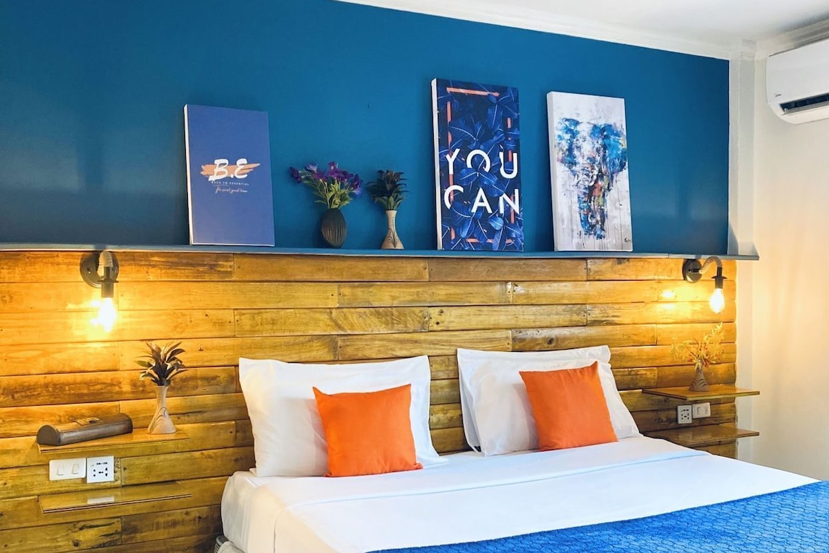 Habitación de hotel con cabecera de madera, cama con ropa de cama blanca y naranja, y una obra de arte en la pared.