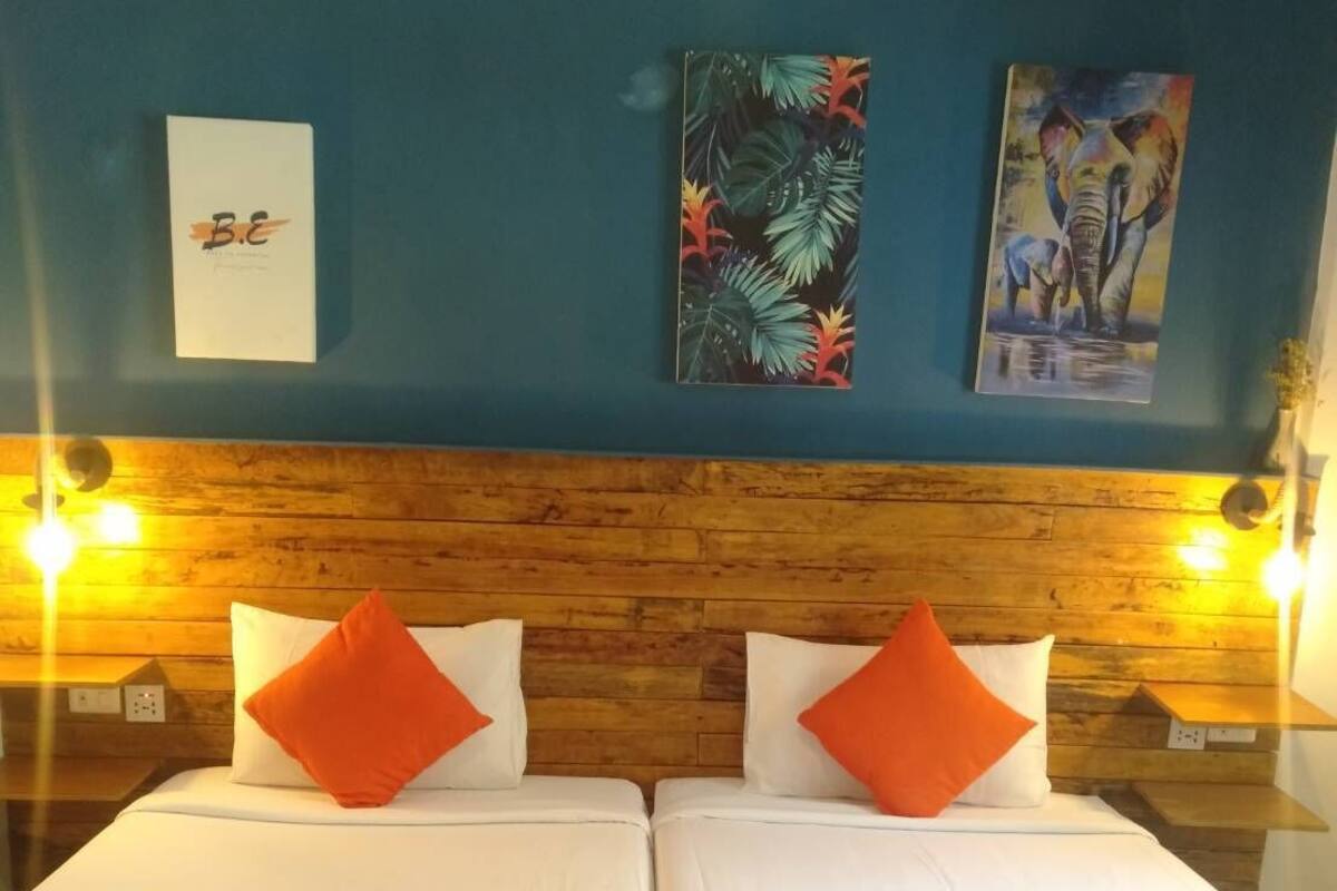 Habitación de hotel con dos camas, cada una con ropa blanca y almohadas naranjas. Dos cuadros colgados en la pared, uno con motivos de hojas tropicales y otro con un elefante. La habitación tiene cabeceros y mesitas de noche de madera con lámparas.