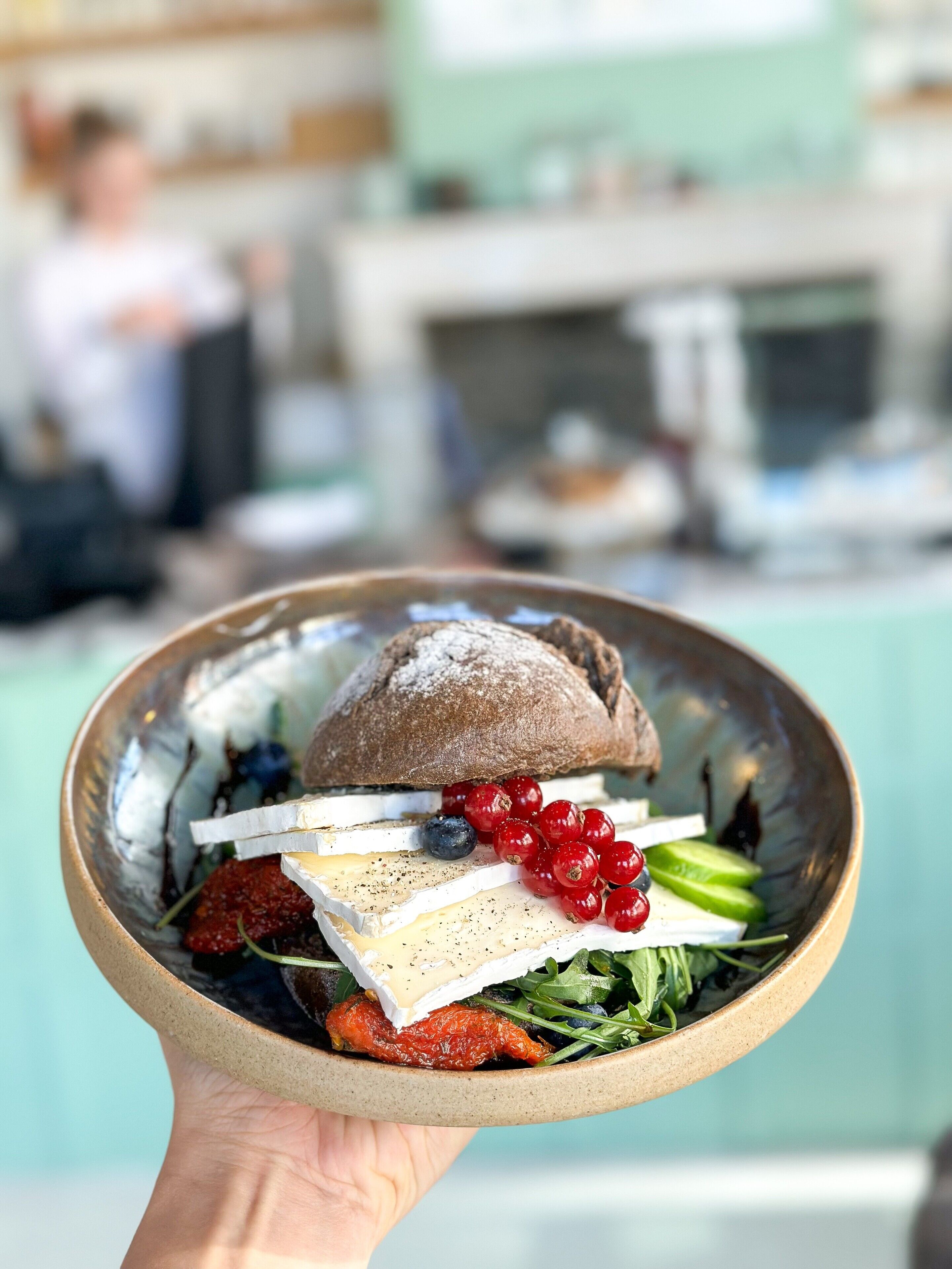 A person holding a bowl with a variety of food items including bread, cheese, berries, and greens.