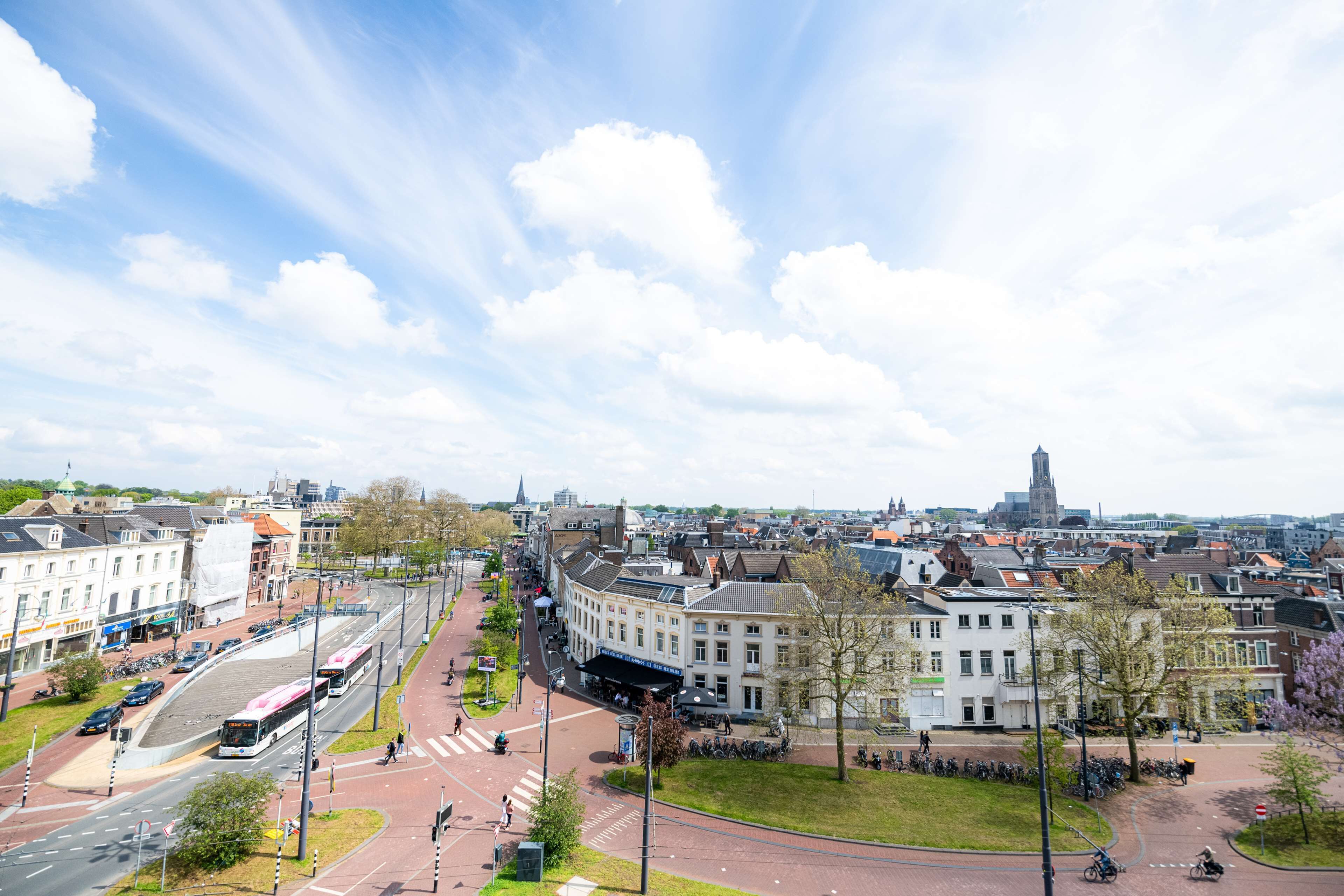 A cityscape with a prominent building with a clock tower, a tram line, and a pedestrian area with trees and benches.