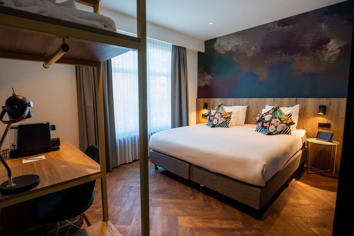 A modern hotel room with a large bed, a desk with a laptop, a small bedside table with a lamp, and a large abstract painting on the wall.