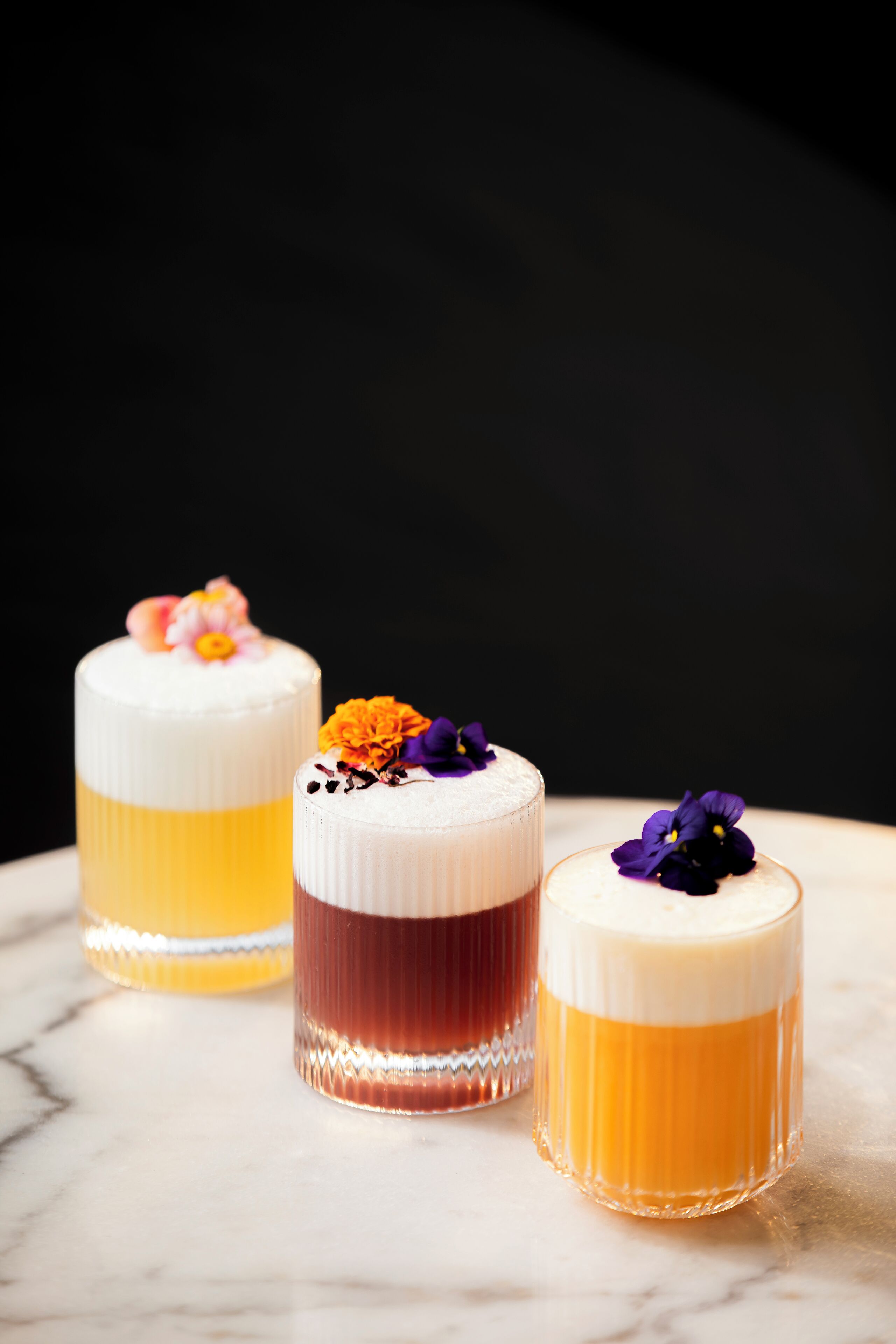 Drei zylindrische Dessertgläser mit verschiedenen Schichten und floralen Verzierungen auf einer Marmorplatte.