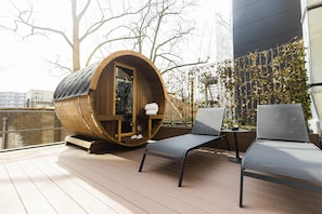 Una sauna de madera con barril, puerta de vidrio, dos sillones reclinables y una mesa en una terraza al aire libre.