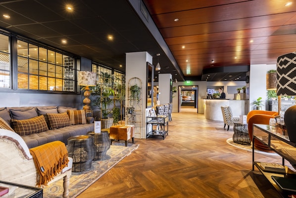 A spacious lobby with a wooden floor, a grey sofa with patterned cushions, a coffee table, and a reception desk. - Hotel Haarhuis, WorldHotels Crafted (Arnhem)