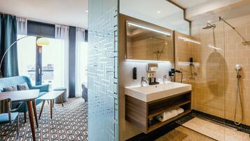 Een moderne hotelkamer met een groot raam, een bank, een eettafel, een badkamer met douche en een spiegel.