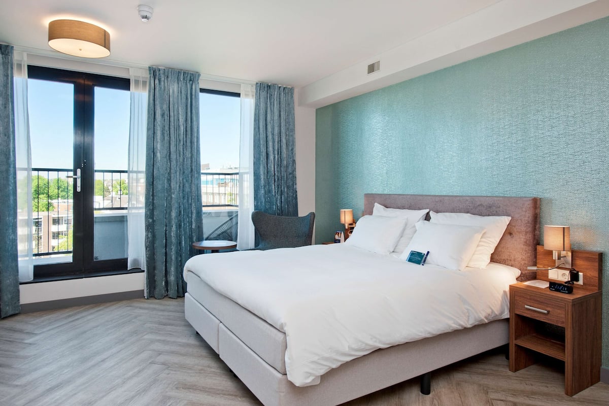 A modern hotel room with a large bed, a wooden nightstand, a chair, and a view of the city through large windows.