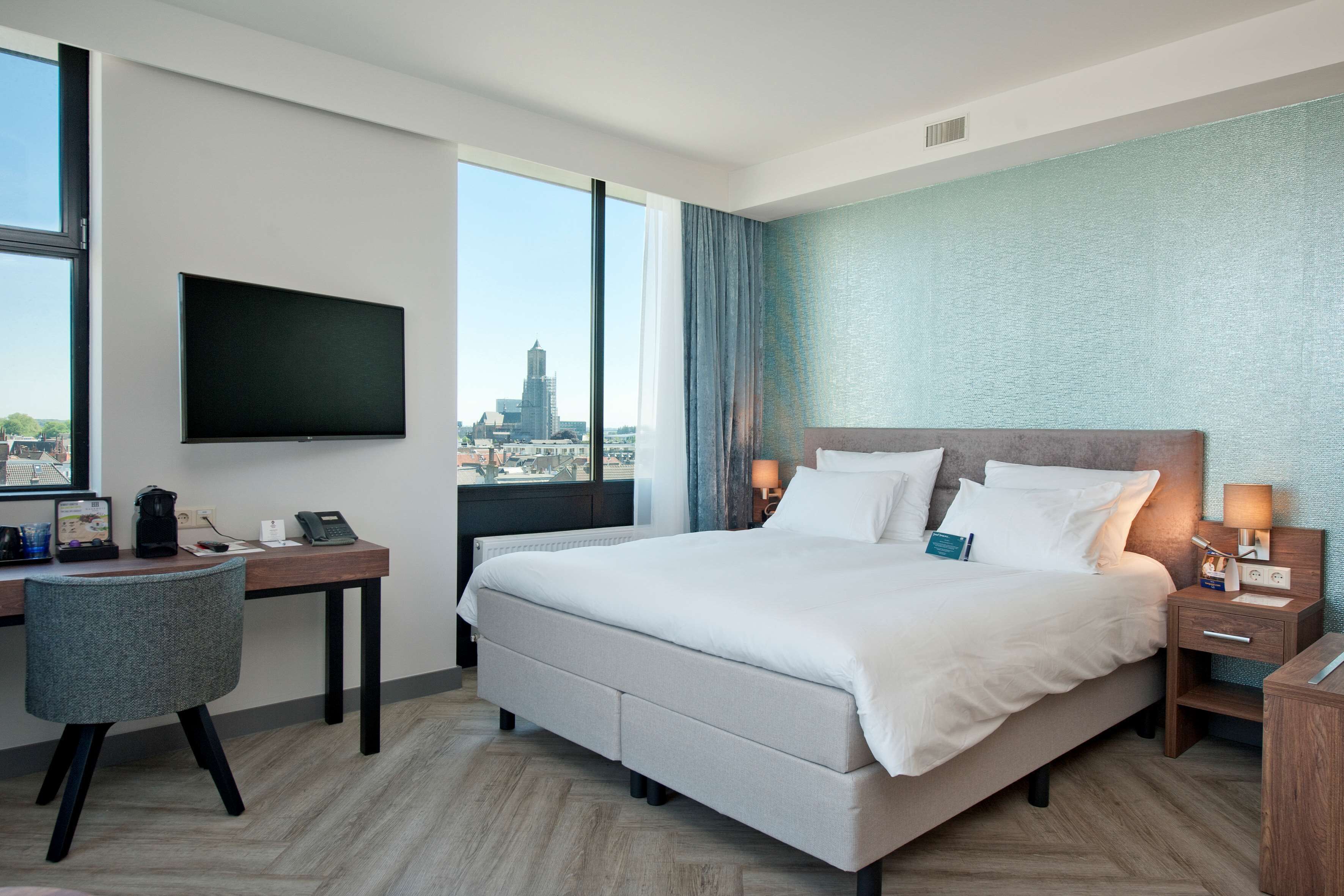 A modern hotel room with a large bed, a desk, a chair, a TV mounted on the wall, and a view of the cityscape.