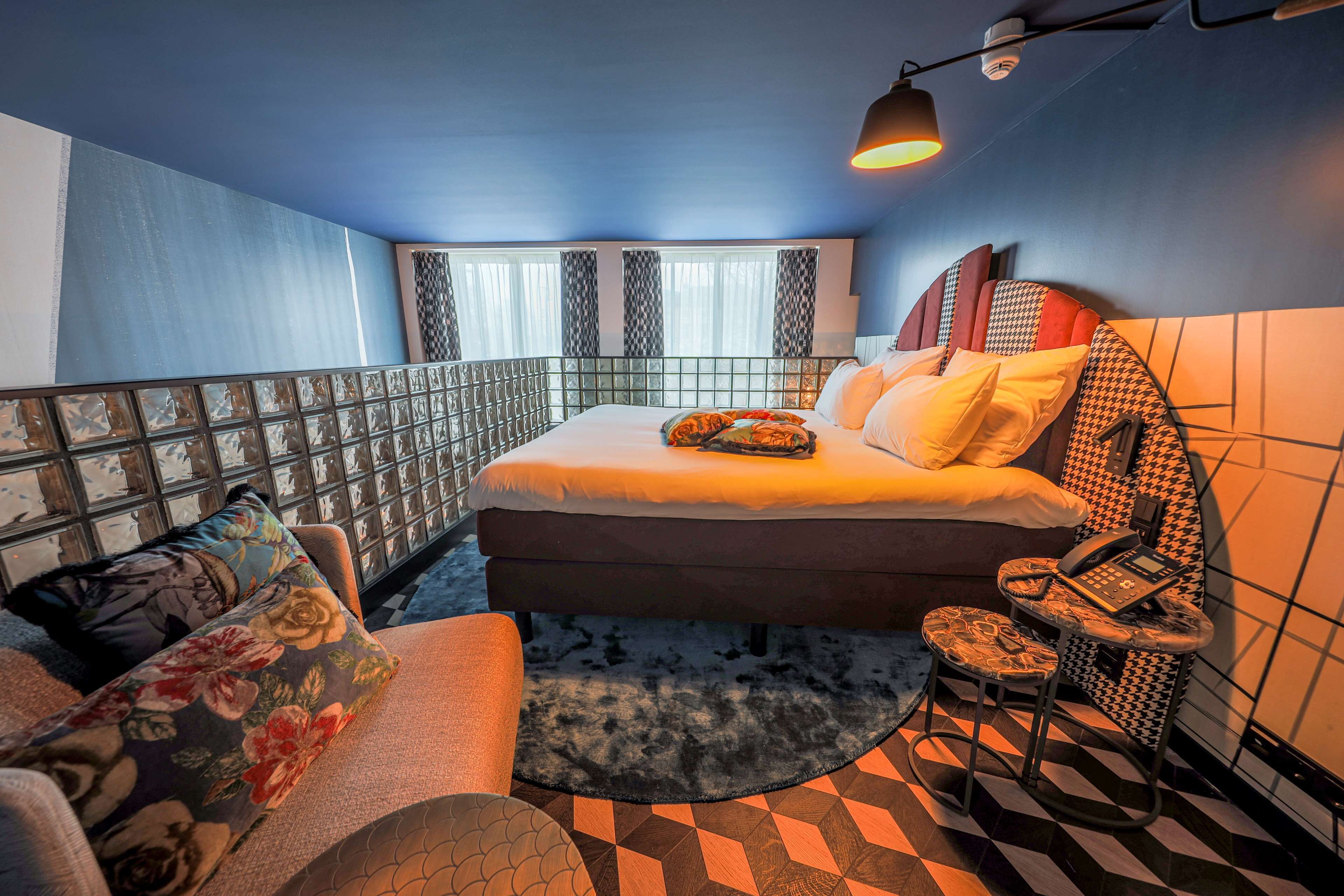 A modern hotel room with a bed, a small table with a telephone, a chair, a sofa, and a patterned rug.