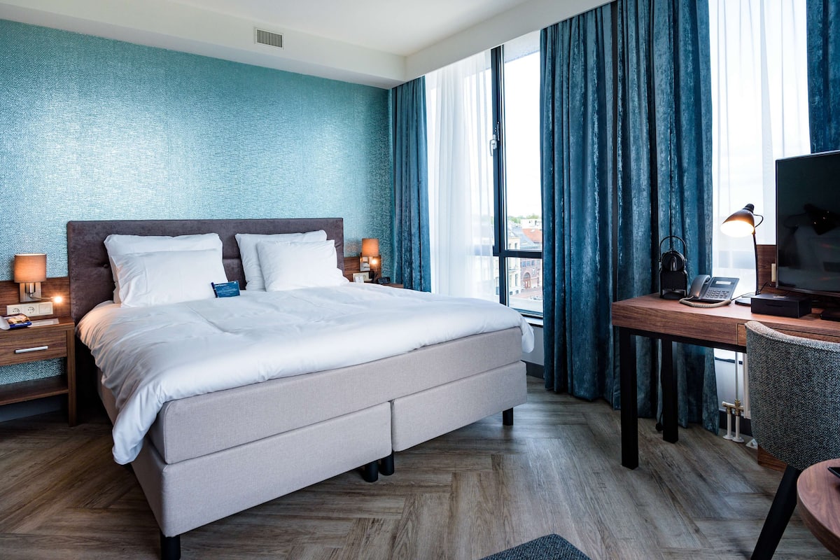 A modern hotel room with a large bed, a desk with a computer and phone, and a view of the city through large windows.