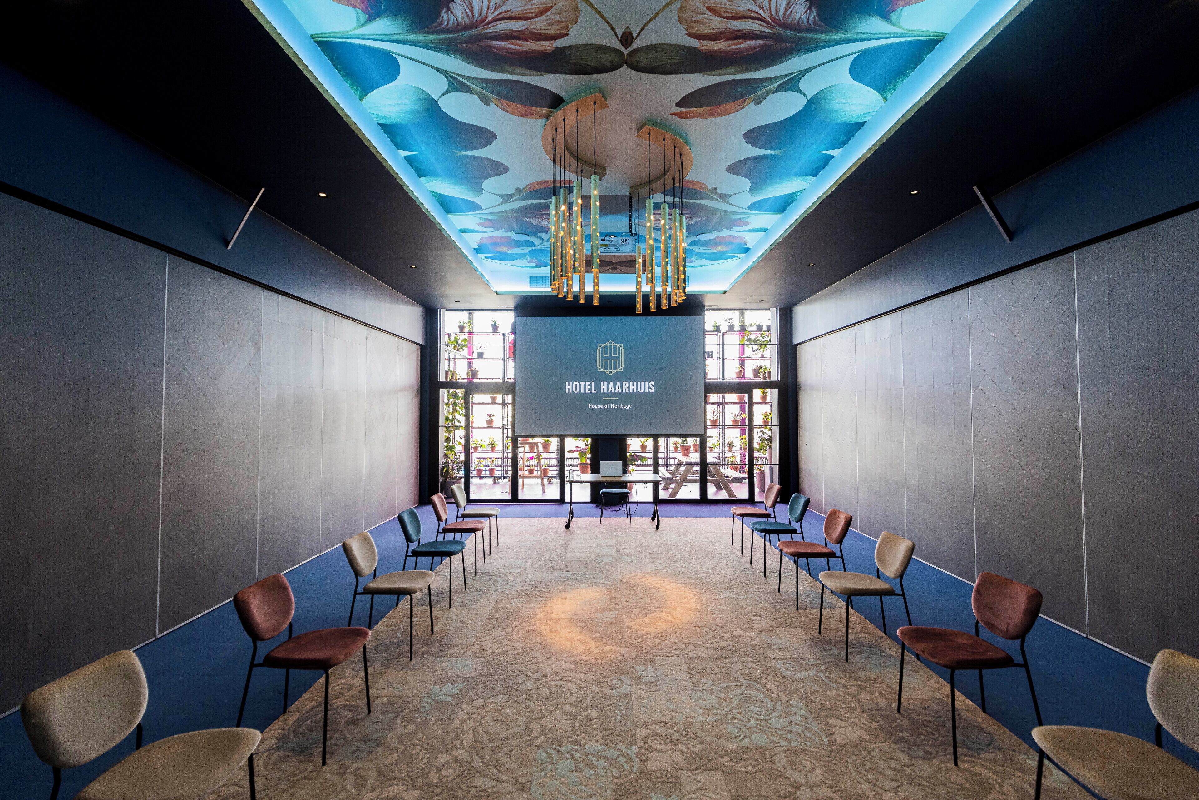 Ein Konferenzraum mit einer großen Leinwand, auf der 'Hotel Haarhuis House of Heritage' steht, mit Stuhlreihen und einem gemusterten Teppich.