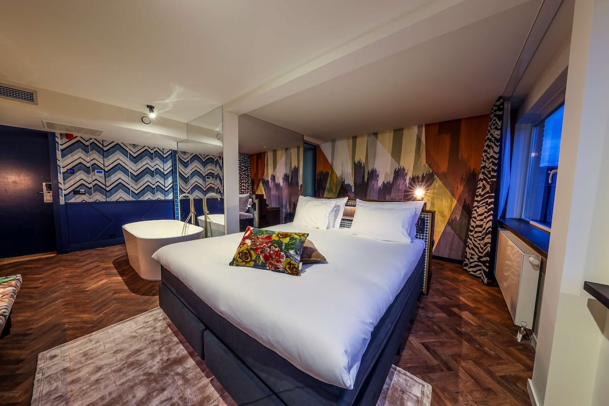A modern hotel room with a large bed, a sitting area, a bathroom, and a cityscape mural on the wall.