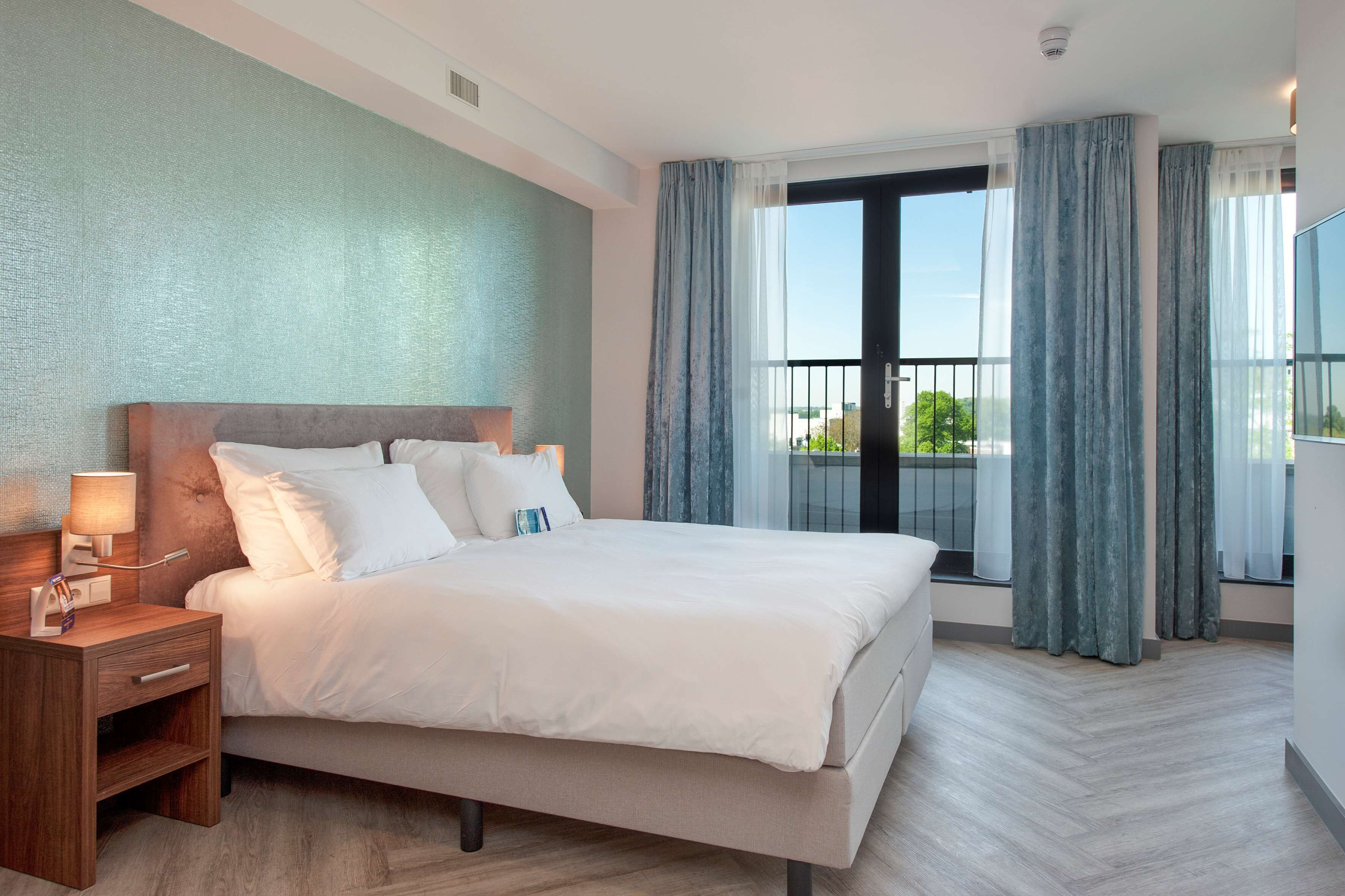 A modern hotel room with a large bed, a bedside table, and a window with curtains.