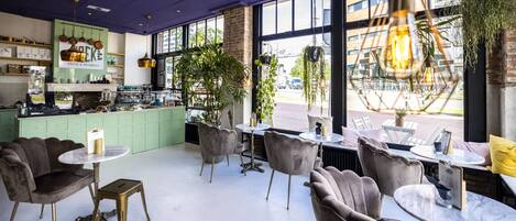 A cafe with marble tables and cushioned chairs, a green counter with kitchen equipment, and large windows with a view of the street.