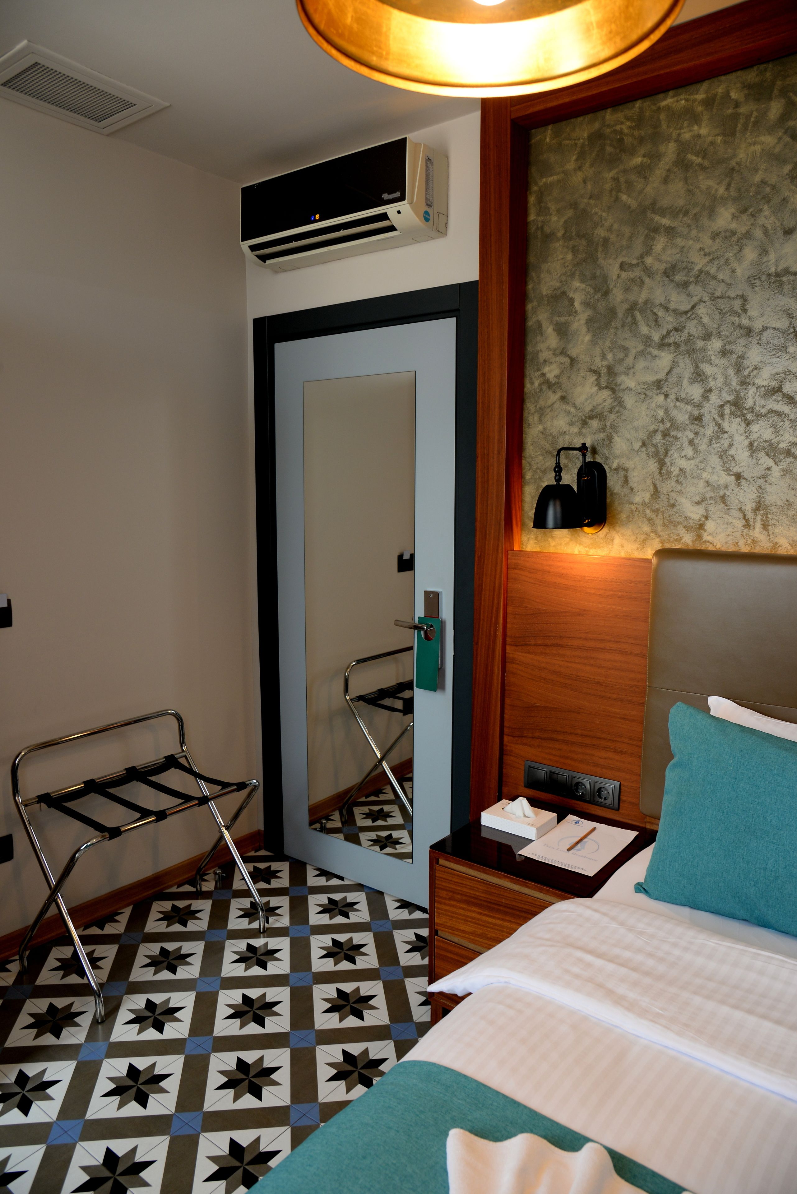 A hotel room with a bed, a bedside table, a mirror, a hanging towel rack, a patterned floor, and an air conditioning unit.
