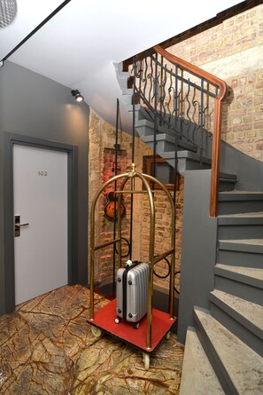 Um quarto com uma mala num carrinho vermelho, um violino encostado a uma parede de tijolos e uma escada com corrimão de metal.