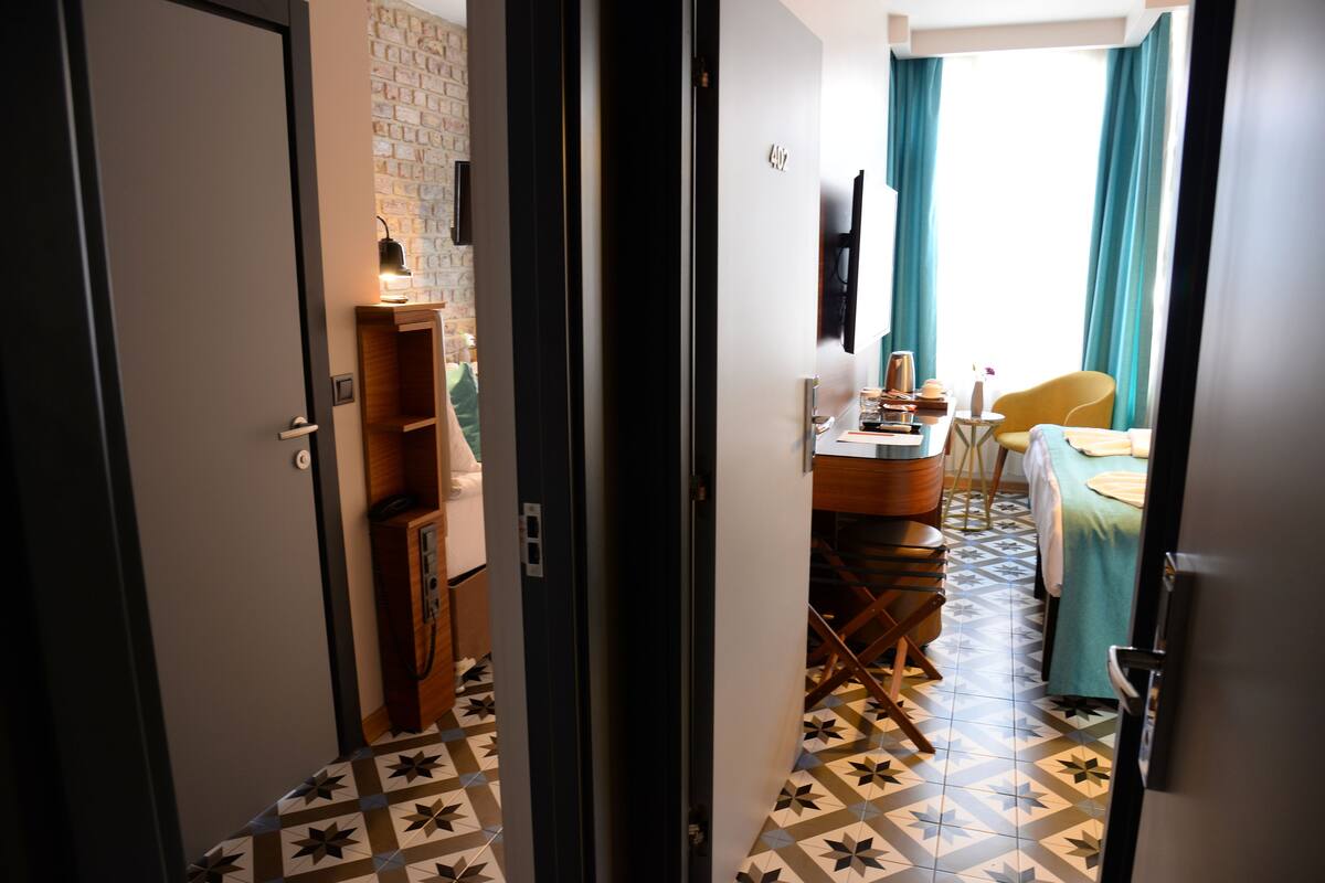 Un couloir étroit mène à une pièce où se trouvent un lit, un bureau et une chaise. Un meuble en bois et une lampe sont fixés au mur.
