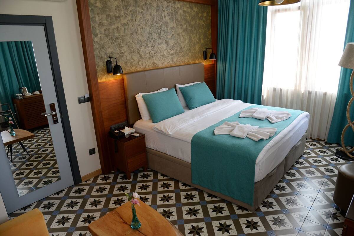Une chambre d’hôtel comprenant un lit, des tables de chevet, une commode, un miroir, une fenêtre avec des rideaux et un sol à motifs.