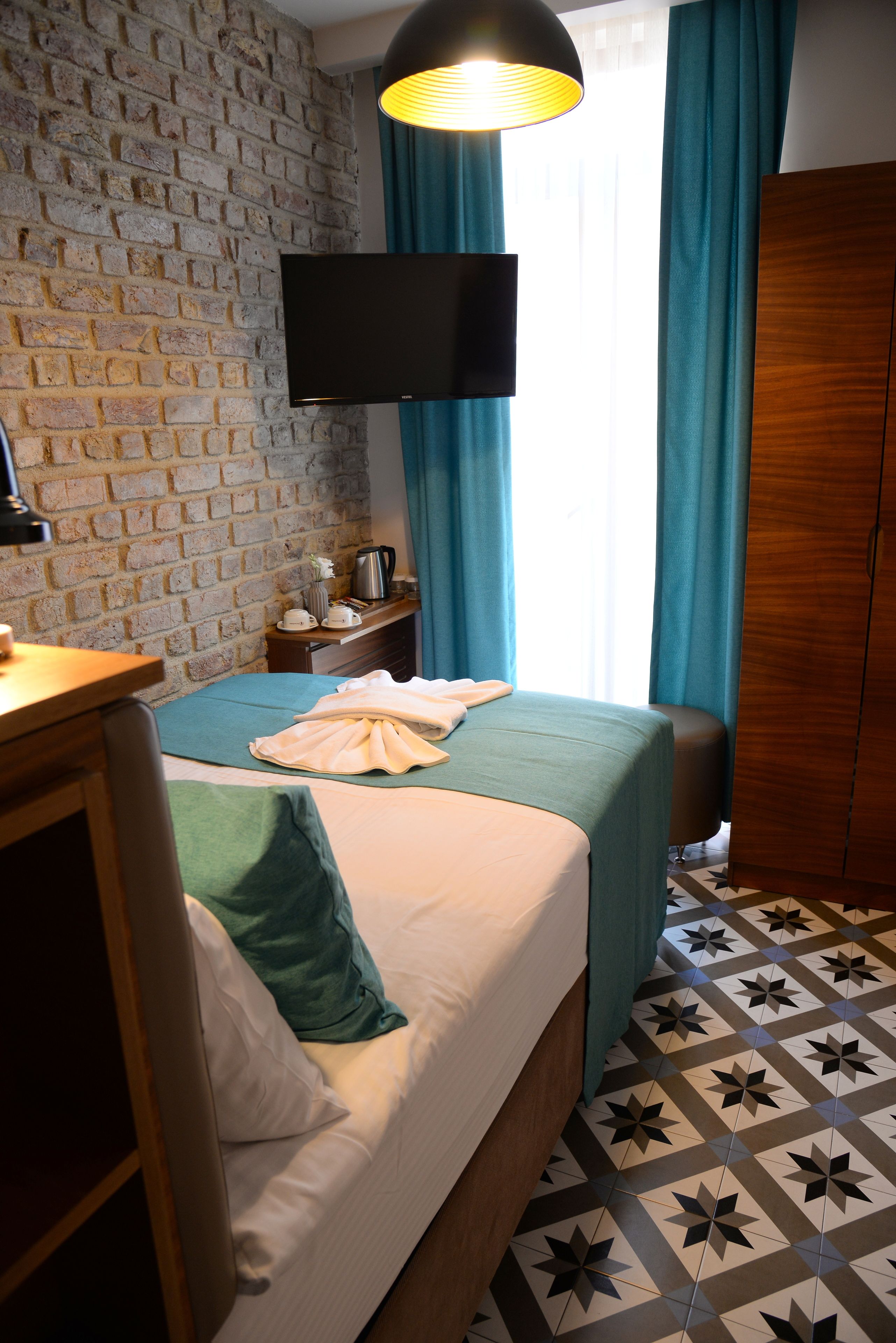A hotel room with a brick wall, a bed with a green bedspread, a wooden wardrobe, a flat-screen TV, and a window with blue curtains.