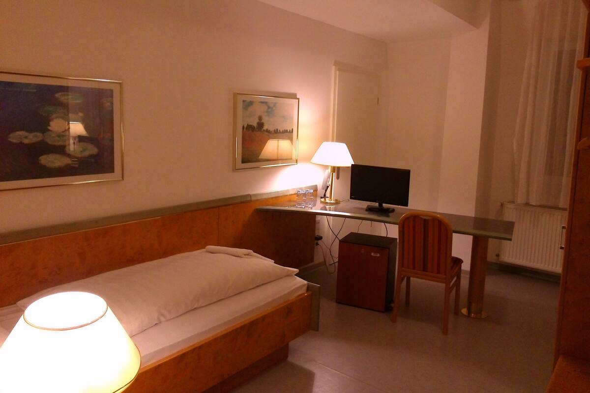 Een hotelkamer met een bed, een bureau met een computer, een stoel, een lamp en een schilderij aan de muur.