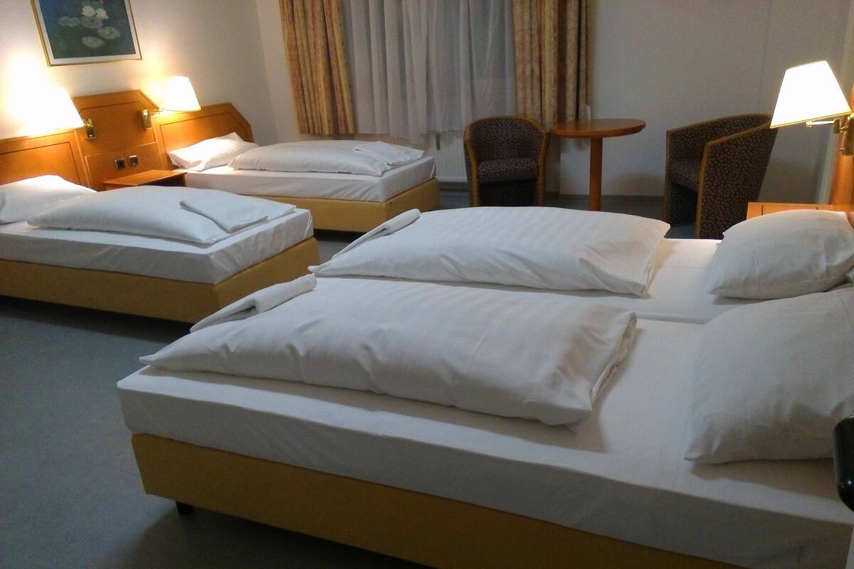 Hotelkamer met drie bedden, elk voorzien van witte lakens en kussens. Twee nachtlampen, een klein rond tafeltje en een stoel.
