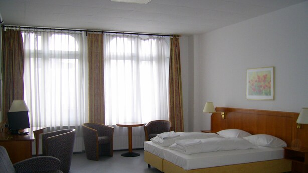 Une chambre d’hôtel comprenant un lit, deux fauteuils, une petite table, un bureau avec une lampe et une fenêtre avec des rideaux.