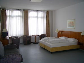 A hotel room with a bed, two armchairs, a small table, a desk with a lamp, and a window with curtains. - Willy Hotel Frankfurt (Frankfurt)