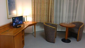 Une chambre d’hôtel avec un bureau, deux fauteuils à motifs, une petite table avec deux verres, une télévision et des rideaux.