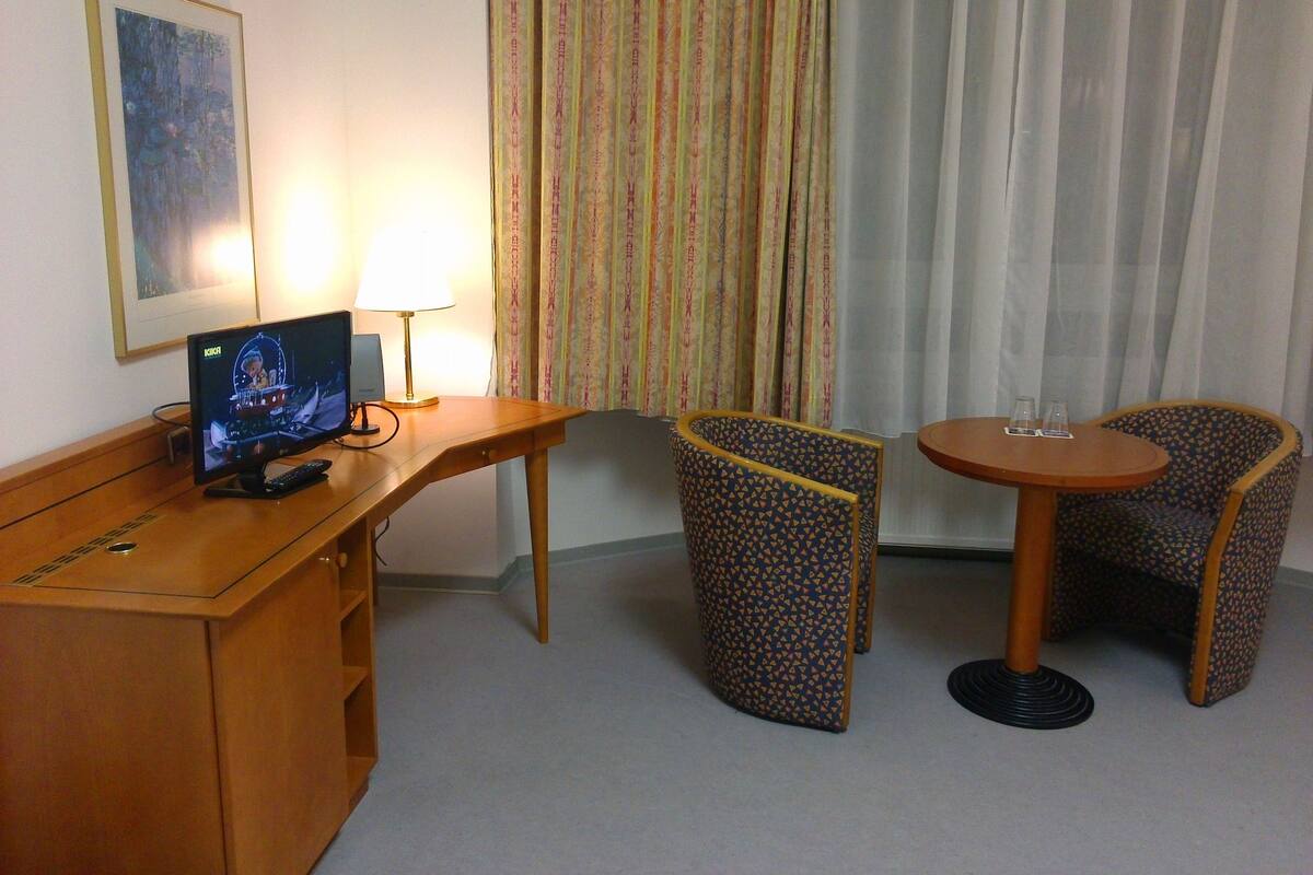 Hotelkamer met een bureau, twee gestreepten stoelen, een tafeltje met twee glaasjes, een televisie en gordijnen.