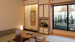 A traditional Japanese room with tatami flooring, a low table with cushions, a tea set, a calligraphy scroll, and a view of a garden through a sliding door. - The Edo Sakura (Tokyo)