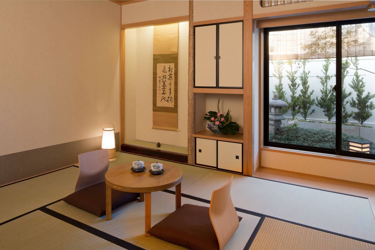Una habitación japonesa tradicional con suelo de tatami, una mesa baja con cojines, un juego de té, un rollo de caligrafía y vistas a un jardín a través de una puerta corredera.