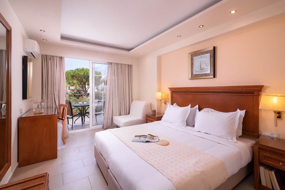 Une chambre d’hôtel avec un grand lit, une tête de lit en bois, deux tables de chevet avec des lampes, une chaise, une petite table avec une lampe et une vue sur l’extérieur grâce à une porte-fenêtre coulissante.