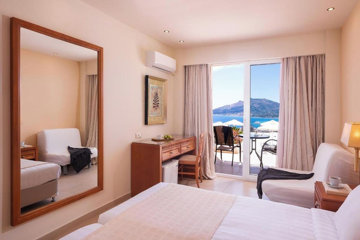 Habitación de hotel con una cama grande, un escritorio, una silla, un aparador y un balcón con vista al mar.