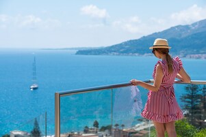 Una donna con un abito e un cappello a righe in piedi accanto alla ringhiera di un balcone, con vista sulla costa e un veliero.
