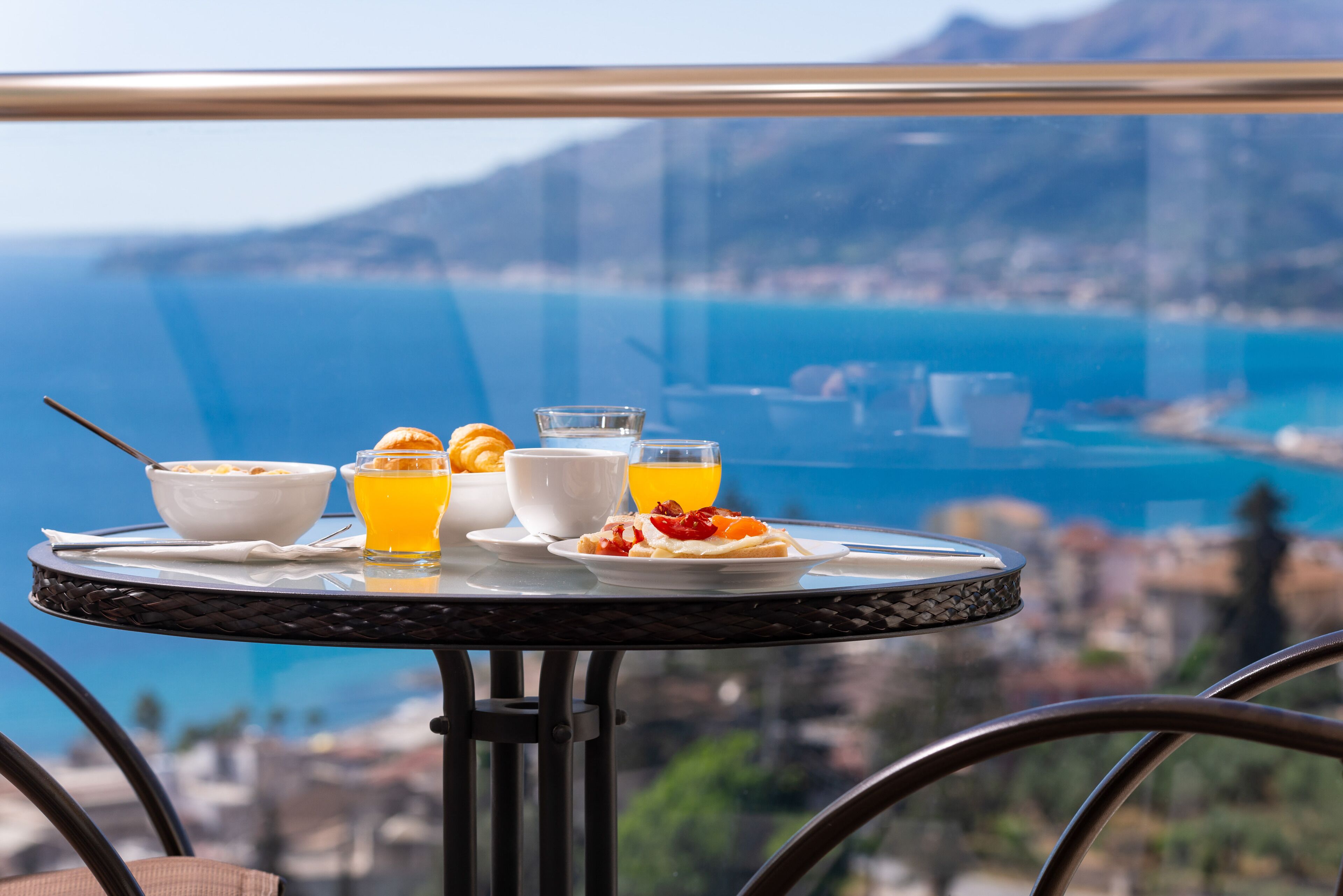 A breakfast table with a bowl of cereal, orange juice, and a plate of food, overlooking a coastal view.