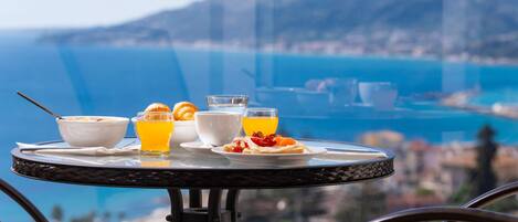 A breakfast table with a bowl of cereal, orange juice, and a plate of food, overlooking a coastal view.