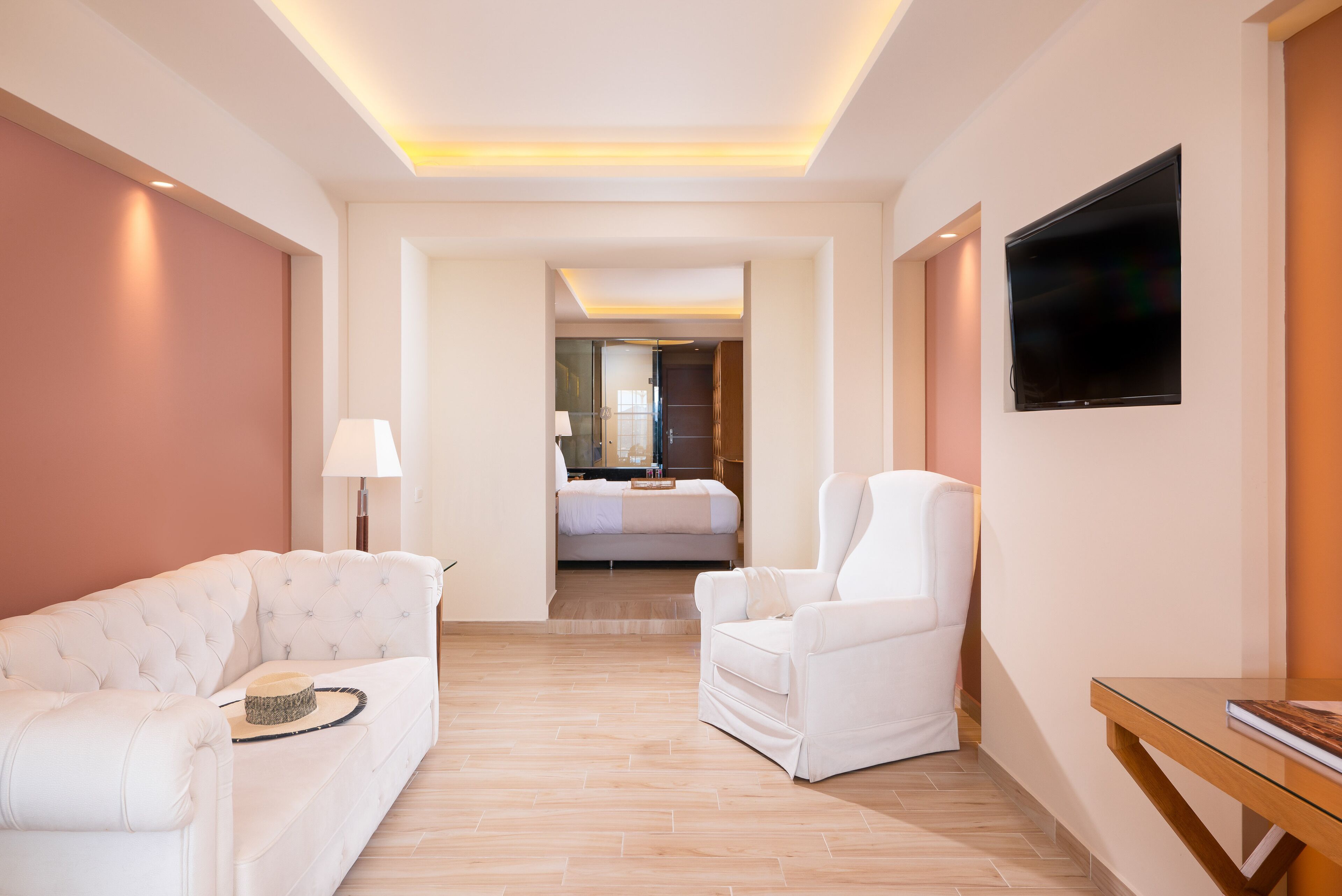 Una habitación de hotel moderna con un sofá blanco, un sillón blanco, un televisor de pantalla plana y una mesita de madera.