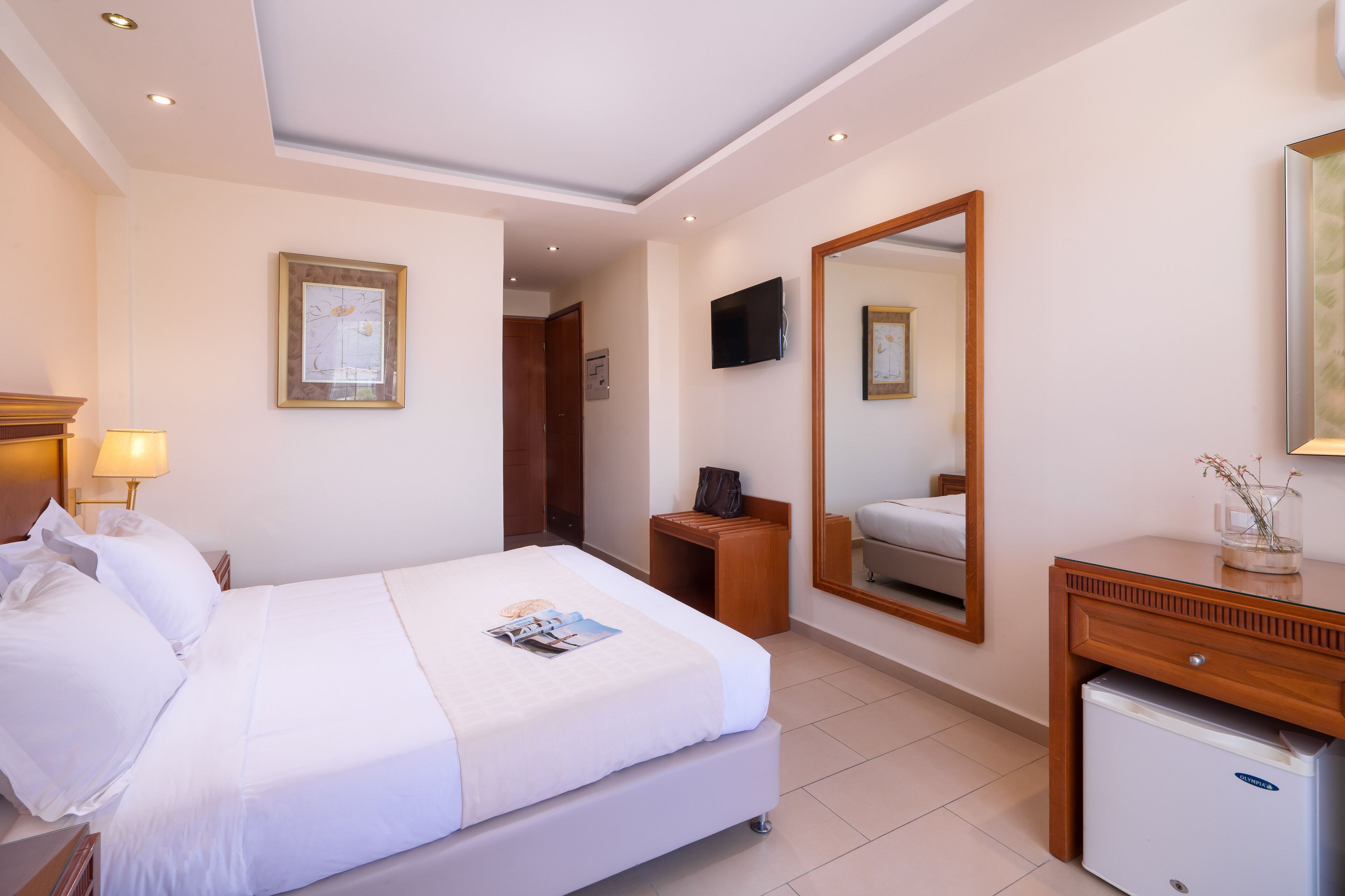 Ein Hotelzimmer mit Bett, Nachttisch, Lampe, Schminktisch, Spiegel, Fernseher und kleinem Kühlschrank.