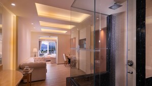 A modern hotel room with a glass-enclosed shower, a sofa, a dining table, and a view of the outdoors.