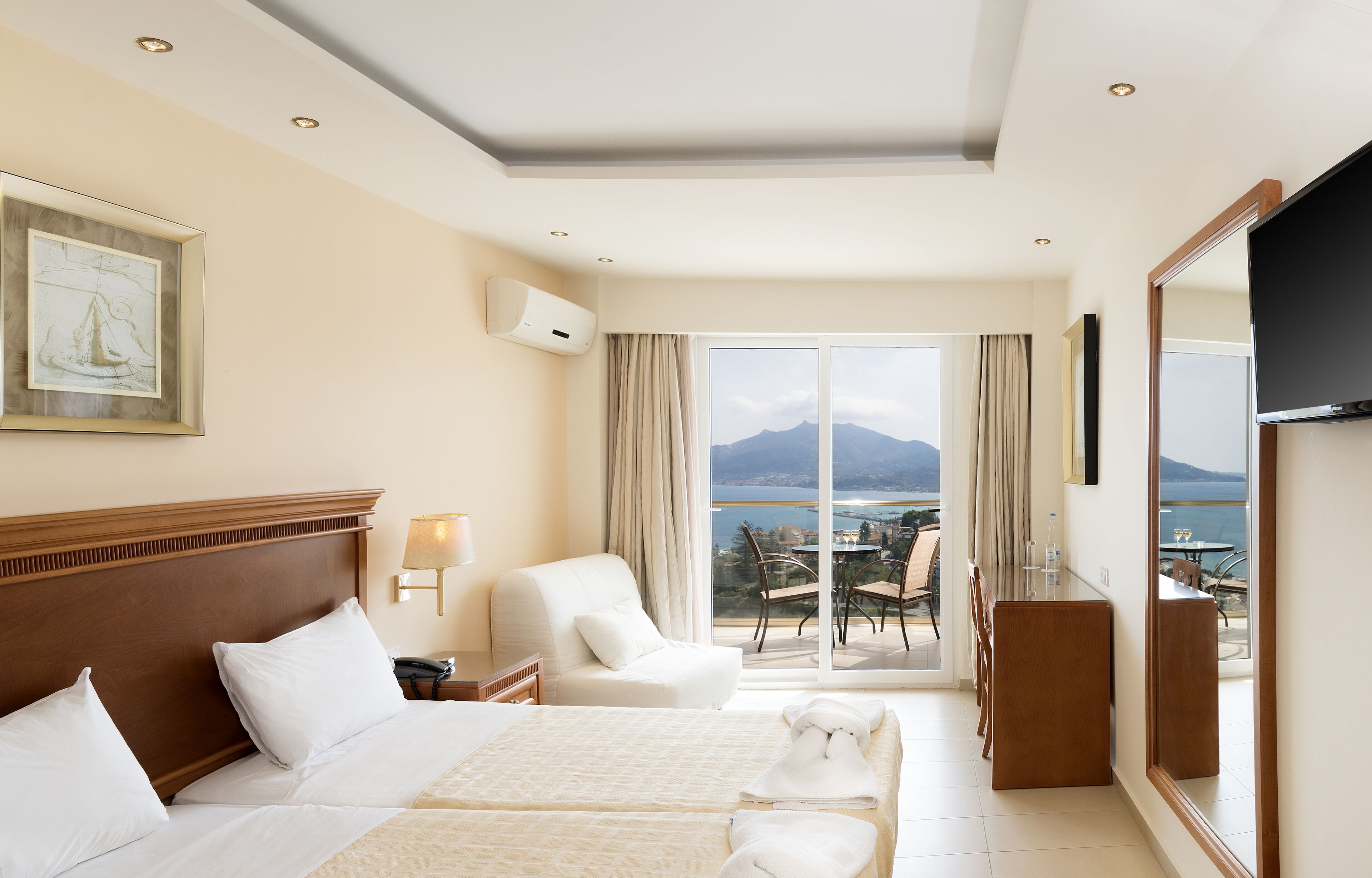 A hotel room with a bed, a desk, a chair, a TV, and a balcony with a view of the sea.