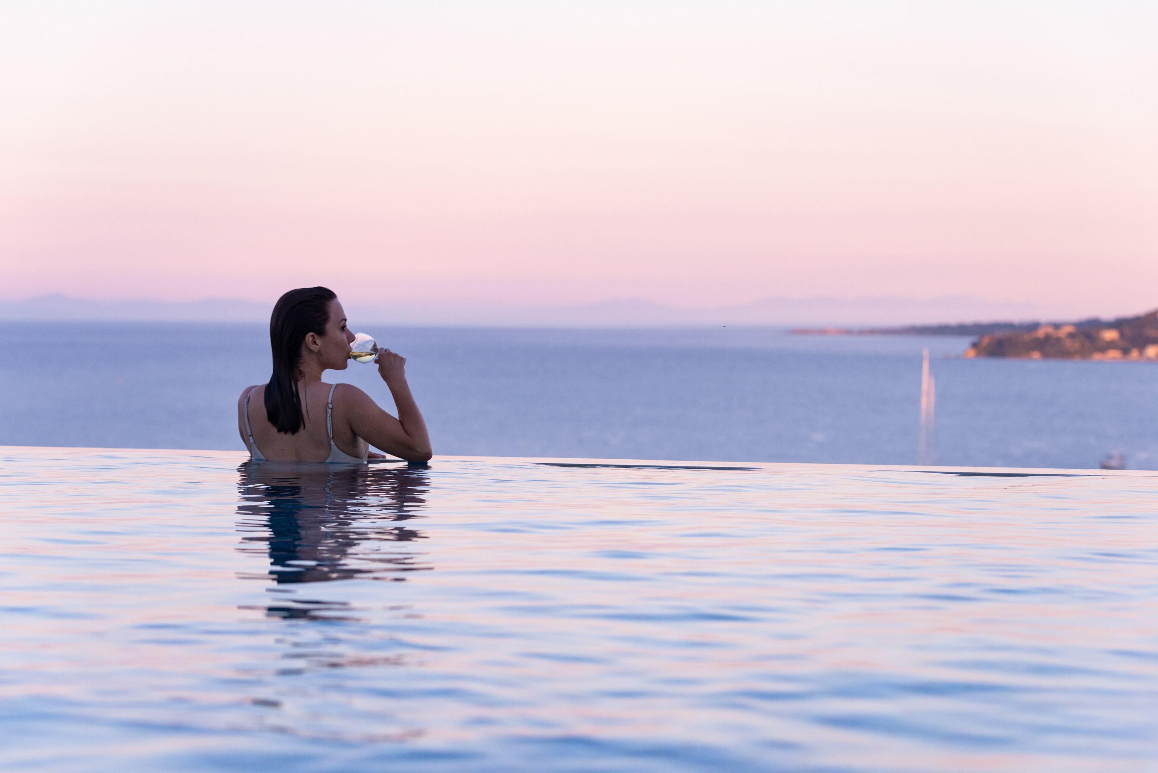 A person relaxing in a clear infinity pool with a view of the sea and distant mountains.