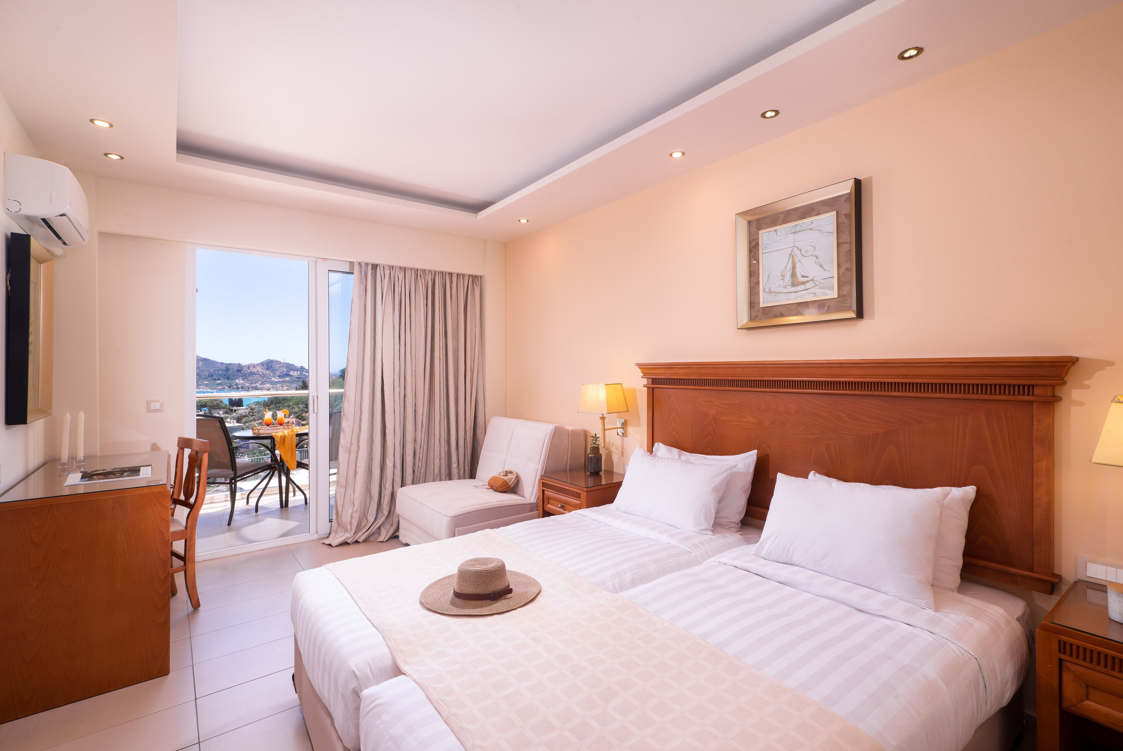 Une chambre d’hôtel avec deux lits, un bureau, une chaise, un balcon avec une table et des chaises, et un tableau au mur.