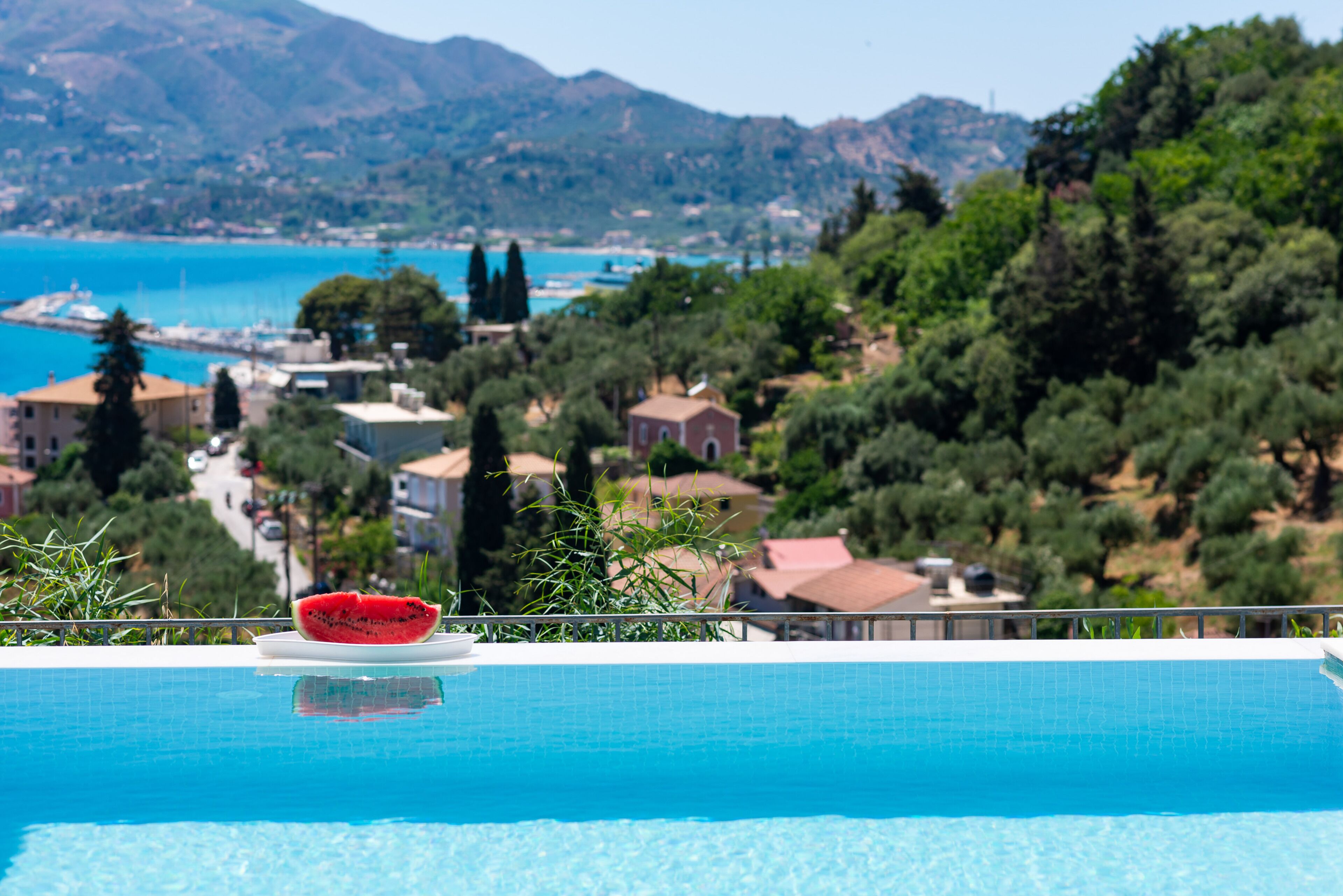 A swimming pool with a slice of watermelon on a white plate, surrounded by greenery and overlooking a coastal town with buildings and mountains in the background.