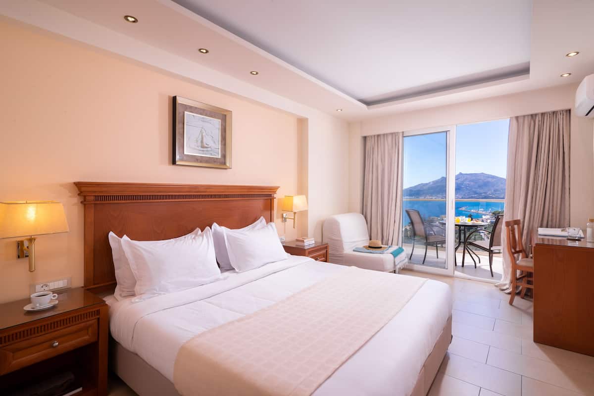 Une chambre d’hôtel avec un grand lit, un bureau, une chaise et une vue sur la mer, accessible par une porte-fenêtre coulissante.