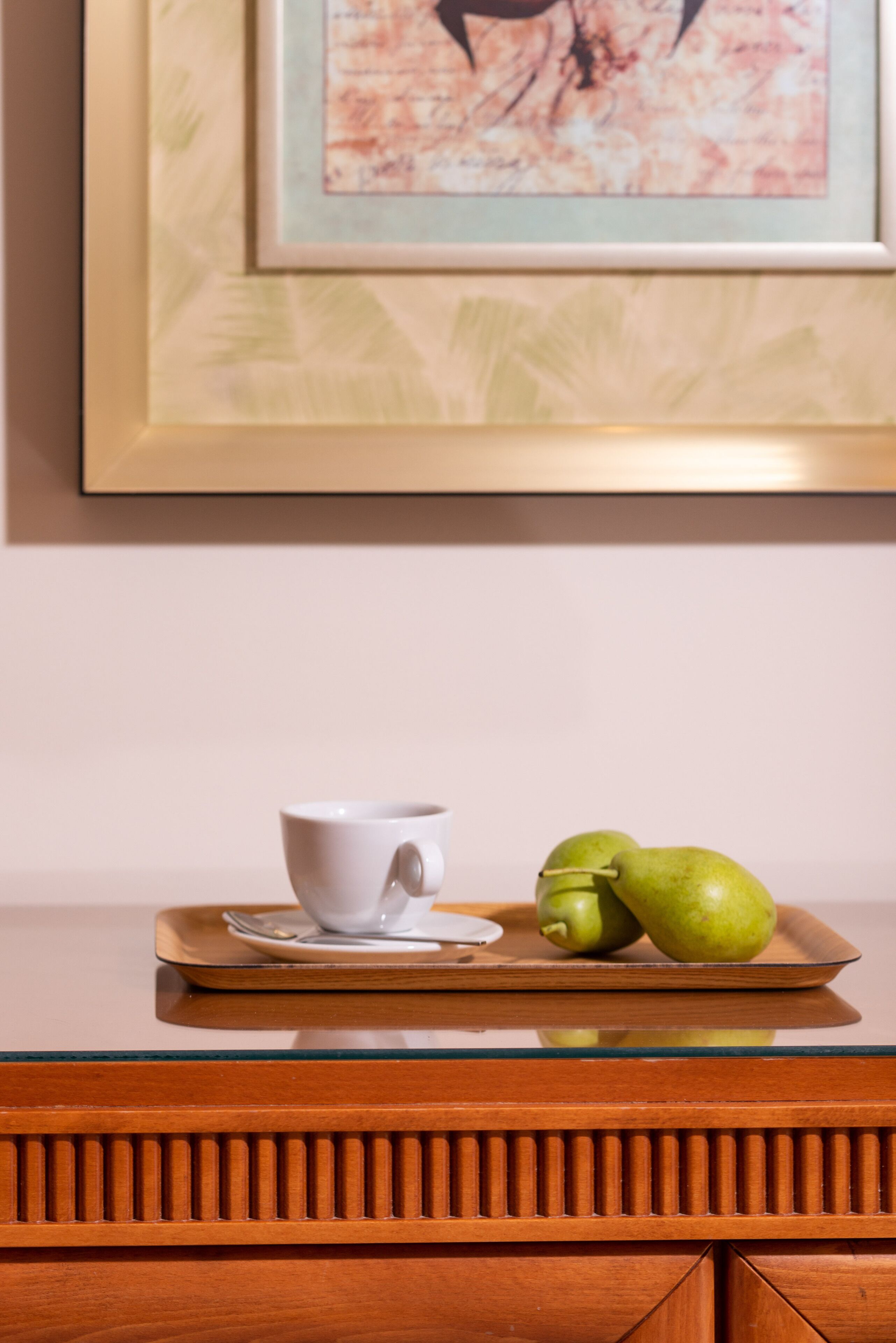 A framed artwork on the wall, a white cup and saucer on a tray, and two green apples on the tray.