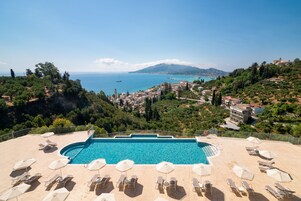 Piscina con lettini e ombrelloni, immersa nel verde e con vista su una località costiera.