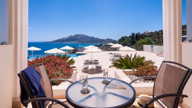 A balcony with a view of the sea, lounge chairs, umbrellas, and a table with glasses.