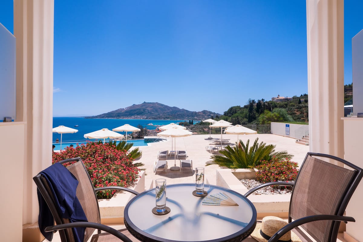 A balcony with a view of the sea, lounge chairs, umbrellas, and a table with glasses.